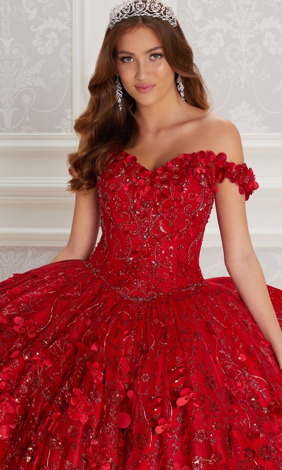 A young woman stands indoors against a patterned white wall, wearing the Flower Quince Dress: Princesa by Ariana Vara PR22036—a sparkling red off-the-shoulder ball gown—paired with a tiara.