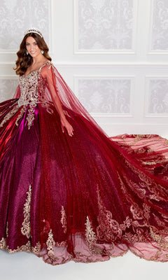 A woman stands against an ornate white wall wearing the Quinceanera Dress PR22141 By Princesa—a sparkling burgundy ball gown with gold embroidery, corset back, long train, and a tiara.