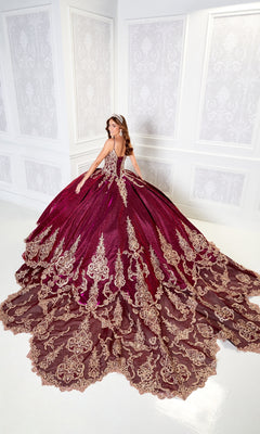 A woman in the Quinceanera Dress PR22141 By Princesa, featuring gold embroidery and a corset back, poses with a tiara in an ornate white room, exuding elegance in her luxurious burgundy ball gown.