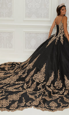 A woman in the Quinceanera Dress PR22141 By Princesa—black with gold embroidery, corset back, and long train—stands in an ornate room, facing slightly away from the camera.
