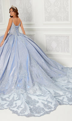 A woman in the Princesa Quinceanera Dress PR22141, featuring a blue ball gown, corset back, long ornate train, and silver embroidery, stands in an elegant white-decorated room, looking over her shoulder.