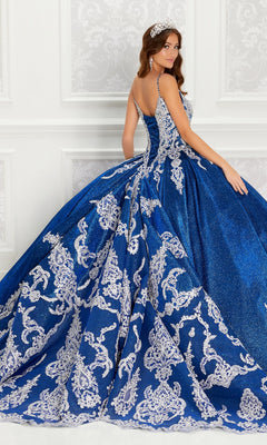 A woman wearing the Quinceanera Dress PR22141 by Princesa, featuring blue and silver embellishments, a corset back, full skirt, and train, stands in an elegant white-decorated room, looking over her shoulder.