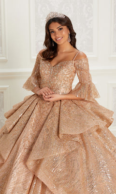 A woman stands indoors wearing the Quinceanera Dress PR22142 By Princesa, featuring sparkling gold fabric, layered ruffles, off-the-shoulder detachable sleeves, a corset back, and accessorized with a tiara and dangling earrings.