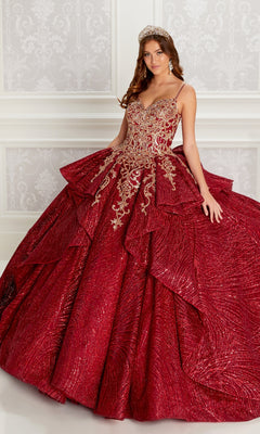 A woman in the Quinceanera Dress PR22142 By Princesa, featuring a sparkling red design with gold embroidery, corset back, detachable sleeves, and a tiara, stands before an ornate white wall radiating elegance.