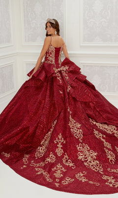 A woman wearing the Quinceanera Dress PR22142 By Princesa, featuring deep red fabric, gold embroidery, and a wide train, stands in a decorated white room with her back turned to show off the elegant corset back.