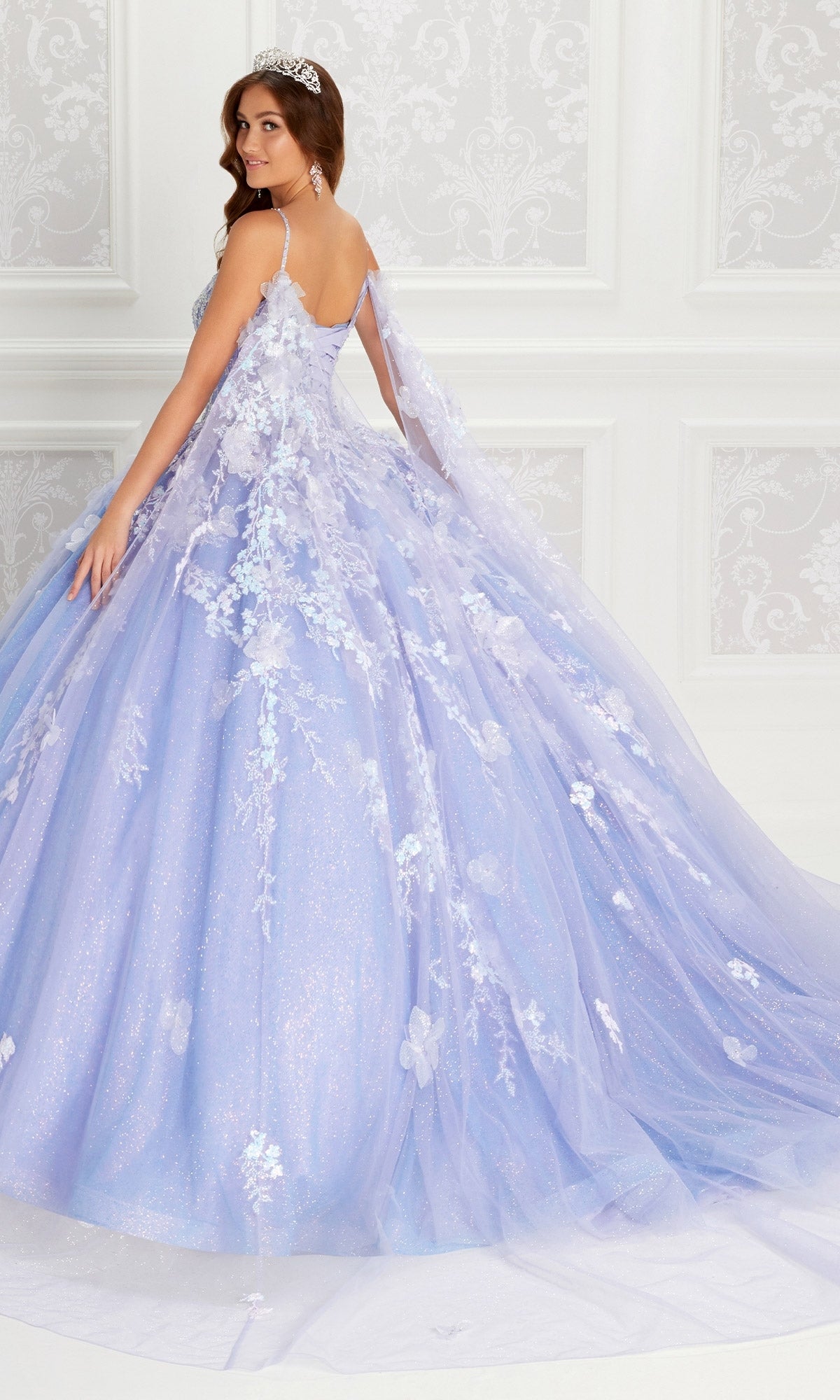 A woman wearing the Quinceanera Dress PR22143 By Princesa, with sparkling lavender fabric, floral appliqués, beaded corset bodice, and a tiara, stands in an ornate white room looking over her shoulder.