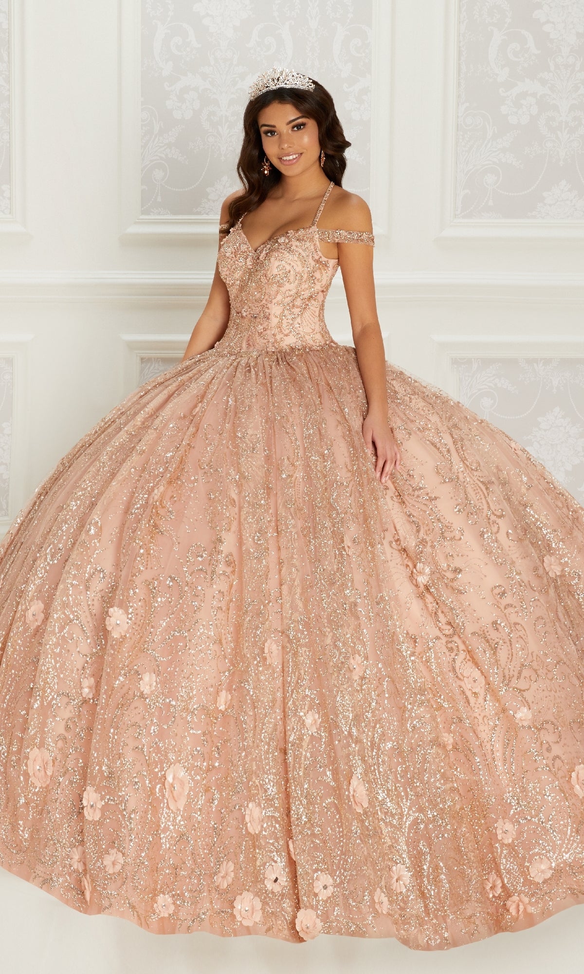 A woman stands in an ornate room with white decorative walls, wearing the sparkling Quinceanera Dress PR22144 by Princesa in peach glitter with detachable straps and a tiara.