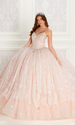 A young woman wears the Quinceanera Dress PR22144 By Princesa in pink glitter with lace, sequin details, corset back, matching tiara, earrings, and necklace, standing before a patterned white wall.