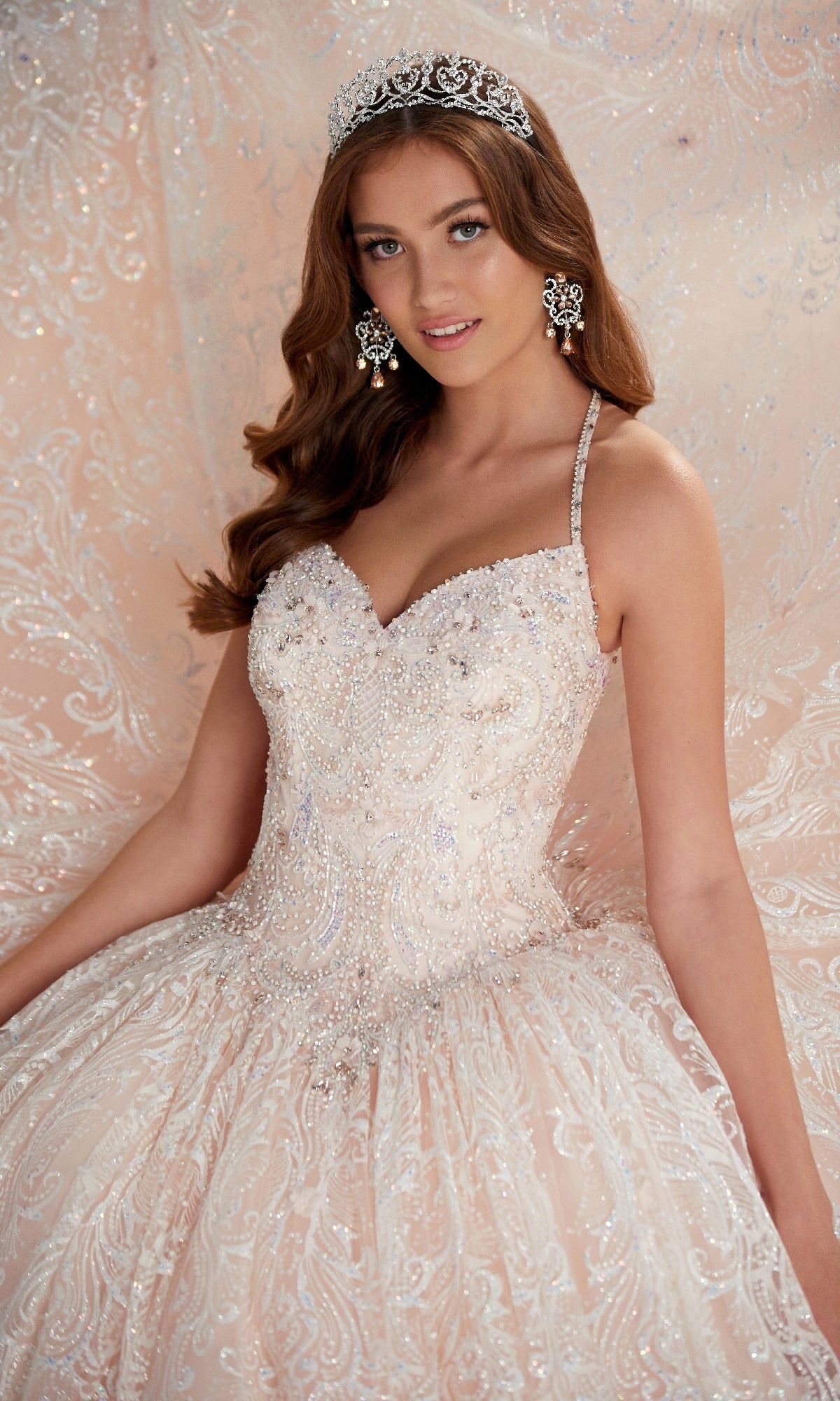 Woman wearing Quinceanera Dress PR22144 By Princesa, featuring a glitter finish, corset back, ornate beading, tiara, and statement earrings, poses in front of a matching intricate fabric backdrop.