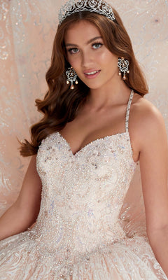 A young woman poses in the Quinceanera Dress PR22144 by Princesa, featuring beaded light-colored glitter fabric, detachable straps, a jeweled tiara, and large drop earrings against an ornate backdrop.
