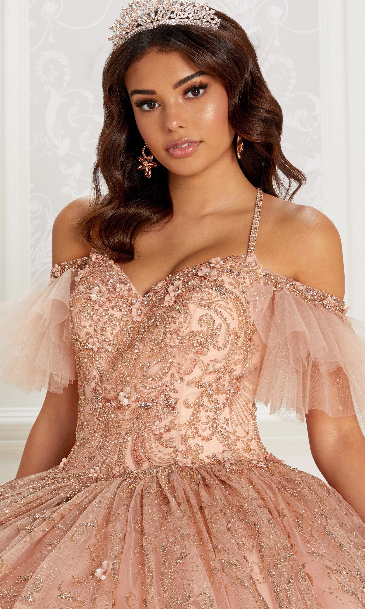 A woman in the Quinceanera Dress PR22144 By Princesa, featuring sparkling peach glitter, off-the-shoulder tulle sleeves, and a corset back with a tiara, stands against a pale decorative background.