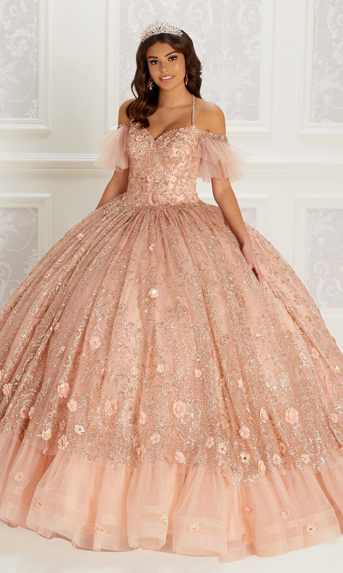 A woman wearing the Quinceanera Dress PR22144 By Princesa, a peach off-the-shoulder glitter gown with floral embroidery, detachable straps, and a tiara stands in an ornate room.