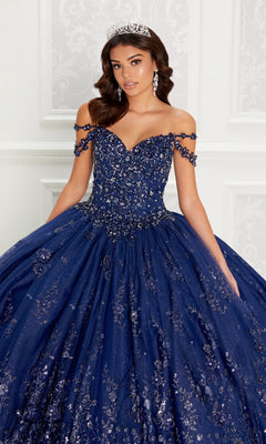 A young woman wears the Quinceanera Dress PR22145 By Princesa, featuring a navy blue off-the-shoulder design with rhinestone embellishments and a corset back, paired with a tiara, against a white ornate background.