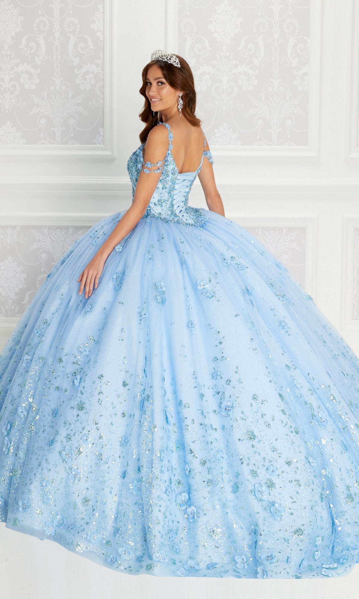A woman stands in an elegant room, looking over her shoulder in the sparkling light blue Quinceanera Dress PR22145 By Princesa, featuring rhinestone embellishments and an off-the-shoulder design, paired with a tiara.