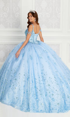 A woman stands in an elegant room, looking over her shoulder in the sparkling light blue Quinceanera Dress PR22145 By Princesa, featuring rhinestone embellishments and an off-the-shoulder design, paired with a tiara.