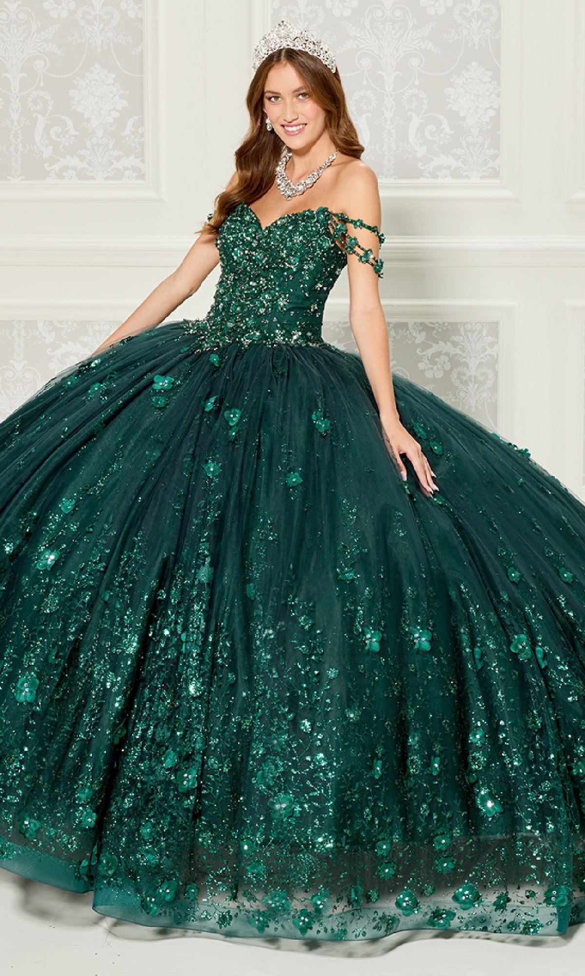 A woman in the Quinceanera Dress PR22145 By Princesa, a sparkling off-the-shoulder gown with rhinestone embellishments and a tiara, stands indoors against a decorative white wall.