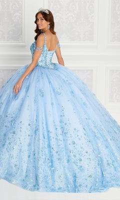 A woman wears the Quinceanera Dress PR22145 By Princesa, a light blue off-the-shoulder gown with floral and rhinestone details, paired with a tiara, as she stands before an ornate white wall.