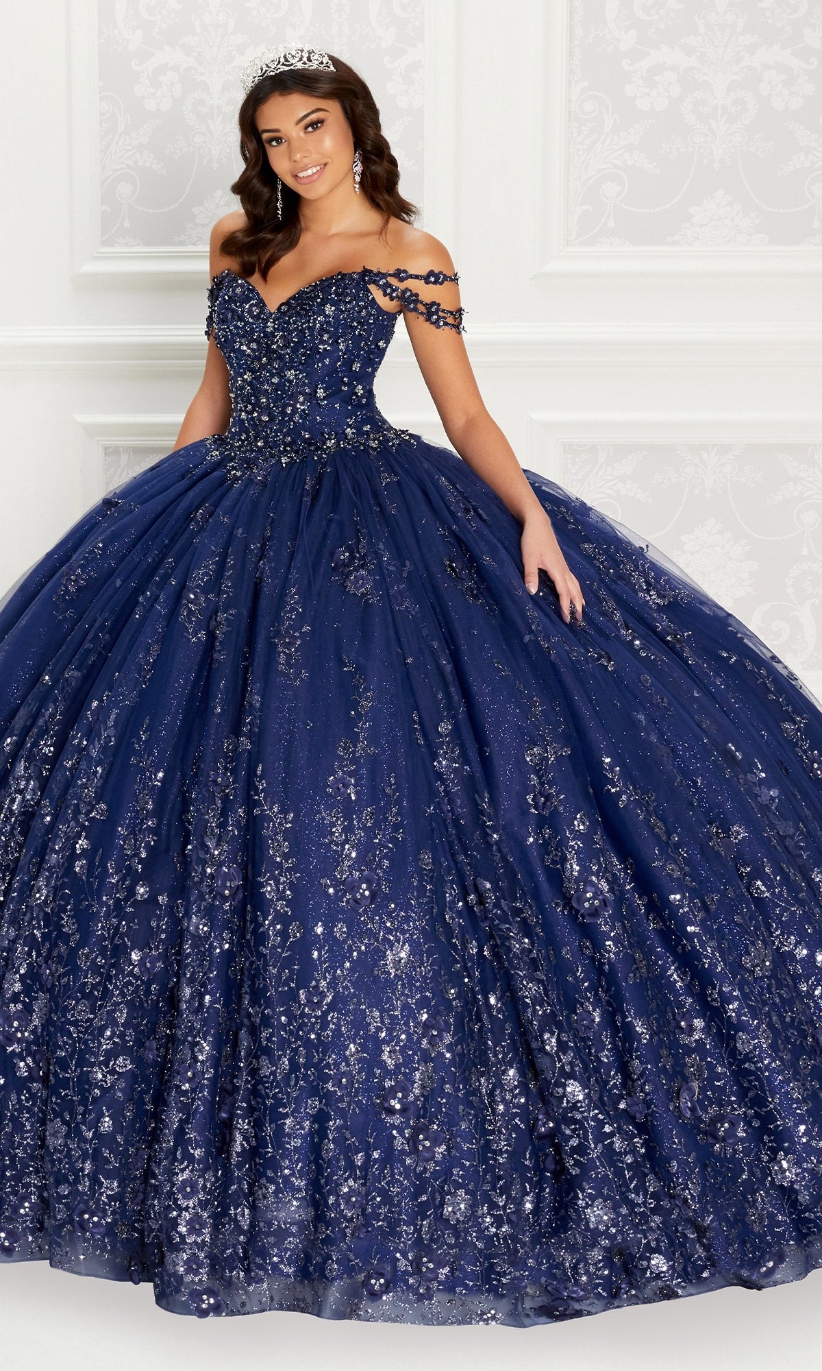 A woman wearing the Quinceanera Dress PR22145 By Princesa, featuring off-the-shoulder rhinestone details and a sparkling tiara, stands before a white ornate background.