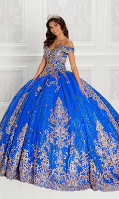 A woman stands gracefully in front of a white paneled wall wearing the Quinceanera Dress PR22148 By Princesa, an ornate off-shoulder royal blue gown with gold lace embroidery, beaded accents, and a sparkling jeweled tiara.
