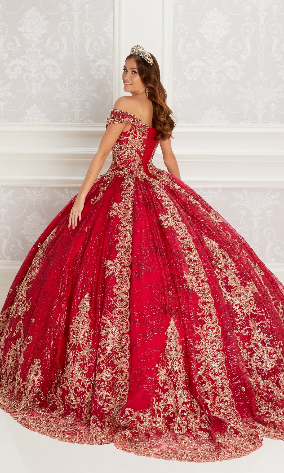 A woman wearing the Quinceanera Dress PR22148 By Princesa, featuring gold and lace embroidery, stands in an ornate white room with a jeweled tiara, smiling over her shoulder.