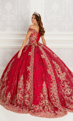 A woman wearing the Quinceanera Dress PR22148 By Princesa, featuring gold and lace embroidery, stands in an ornate white room with a jeweled tiara, smiling over her shoulder.