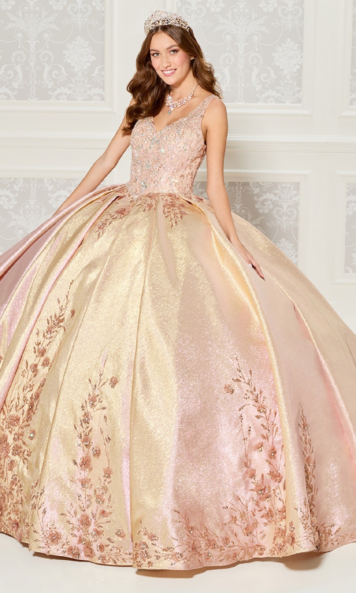 A young woman in a crown wears the Quinceanera Dress PR30081 By Princesa, a sparkling pink and gold metallic jacquard gown with floral embroidery and 3D flowers, as she stands in an ornate room.
