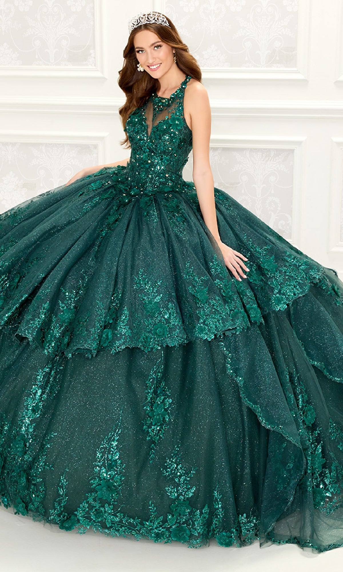A woman wearing the Quinceanera Dress PR30082 By Princesa, a dark green gown with embellishments and an illusion neckline, along with a tiara, stands before a white textured wall.