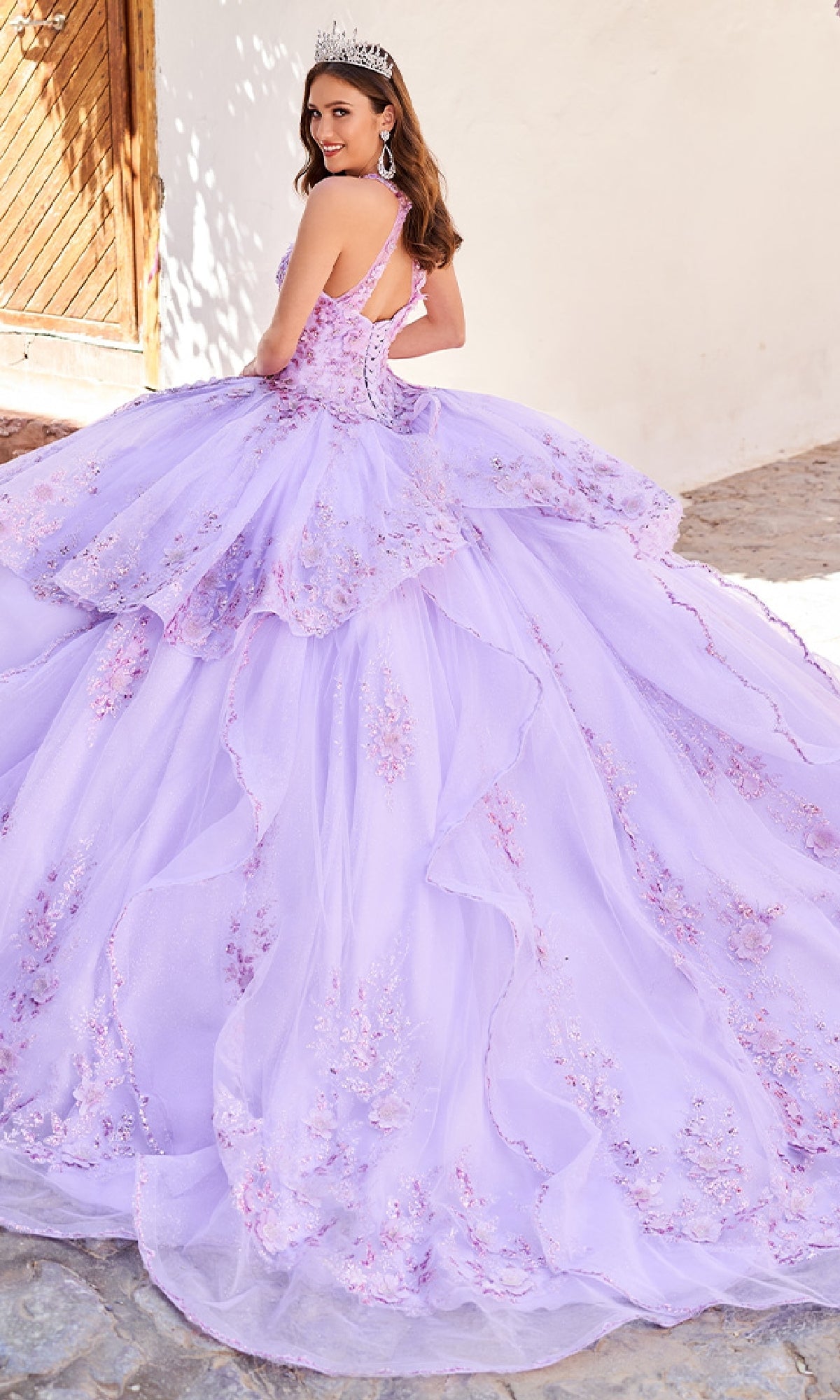 A young woman in the Quinceanera Dress PR30082 By Princesa, featuring embroidery and floral embellishments, stands outdoors in a tiara, looking over her shoulder.