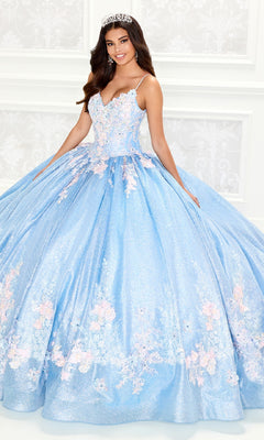 A young woman wearing the Light Blue Quinceanera Dress: Princesa PR30083, featuring a sparkling lace-embroidered corset bodice with floral details and a tiara, smiles as she stands before a white patterned wall.