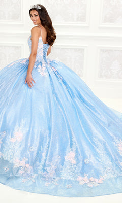 A woman in the Light Blue Quinceanera Dress: Princesa PR30083, featuring pink floral details, lace embroidery, and a corset bodice, stands before an ornate white wall and looks over her shoulder.