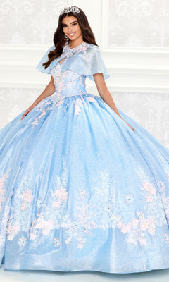 A young woman wearing the Princesa PR30083 Light Blue Quinceanera Dress with a corset bodice, floral details, and a tiara stands in front of a white patterned wall.