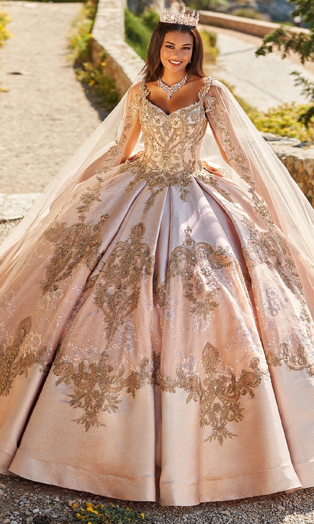 A woman in the Quinceanera Dress PR30085 By Princesa and a jeweled crown stands outdoors, smiling, with a stone wall and greenery behind her.