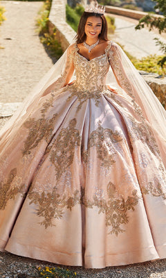 A woman in the Quinceanera Dress PR30085 By Princesa and a jeweled crown stands outdoors, smiling, with a stone wall and greenery behind her.