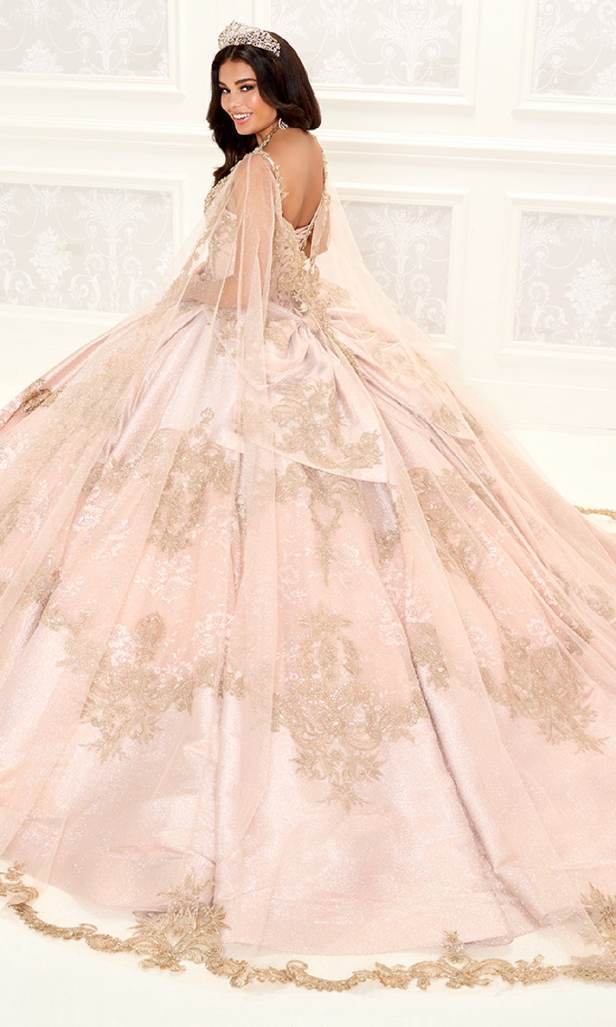 A woman in the Quinceanera Dress PR30085 By Princesa, adorned with gold embroidery and beading, wears a tiara and stands in a white decorated room, looking over her shoulder.