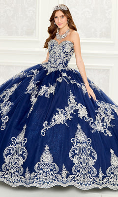A woman poses indoors against a white decorative wall wearing the Quinceanera Dress PR30086 By Princesa, a navy blue ball gown with silver embroidery, a sheer cape, and accessorized with a jeweled tiara and matching necklace.