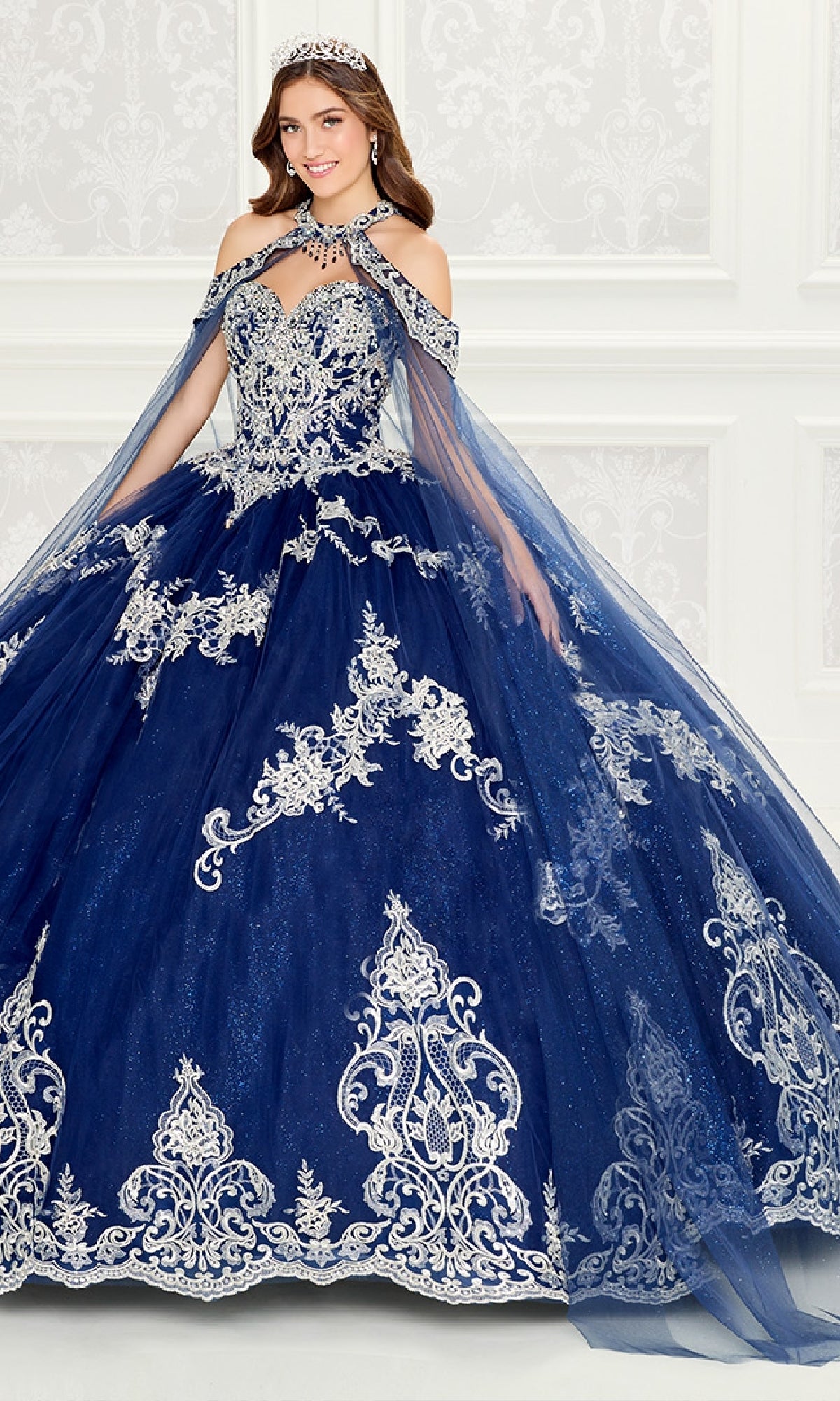 A woman wearing the Quinceanera Dress PR30086 By Princesa, featuring ornate navy blue fabric and silver embroidered lace with a tiara, stands in front of a decorative white wall.