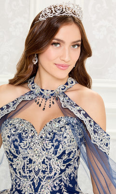 A woman wears the Quinceanera Dress PR30086 By Princesa, a blue and silver gown with intricate embroidery, jeweled accents, a flowing sheer cape, and a sparkling silver tiara.