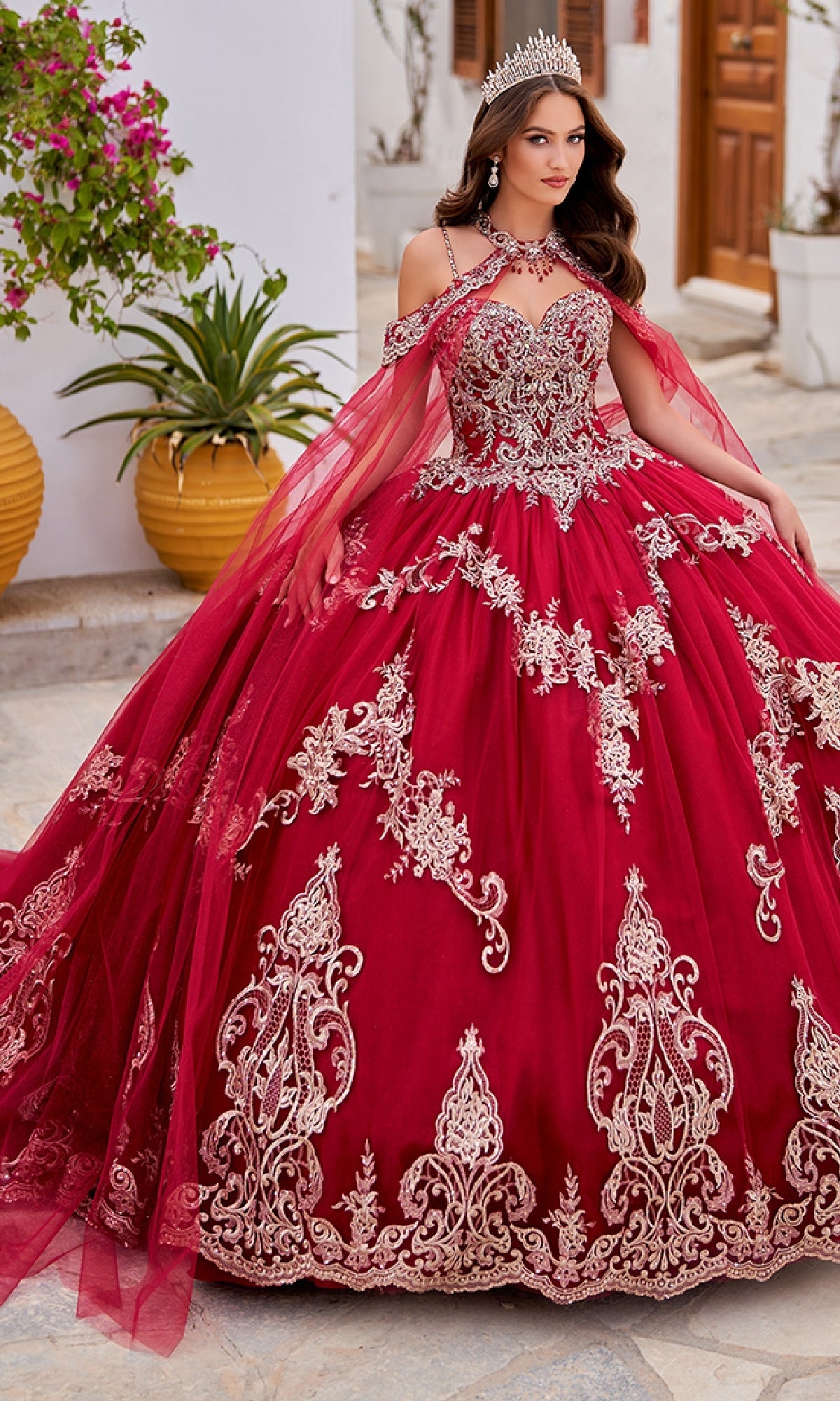 A woman wearing the Quinceanera Dress PR30086 By Princesa, featuring red fabric with gold embroidery and a tiara, stands outdoors on a stone path surrounded by plants and buildings.