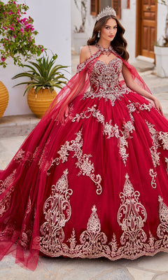 A woman wearing the Quinceanera Dress PR30086 By Princesa, featuring red fabric with gold embroidery and a tiara, stands outdoors on a stone path surrounded by plants and buildings.