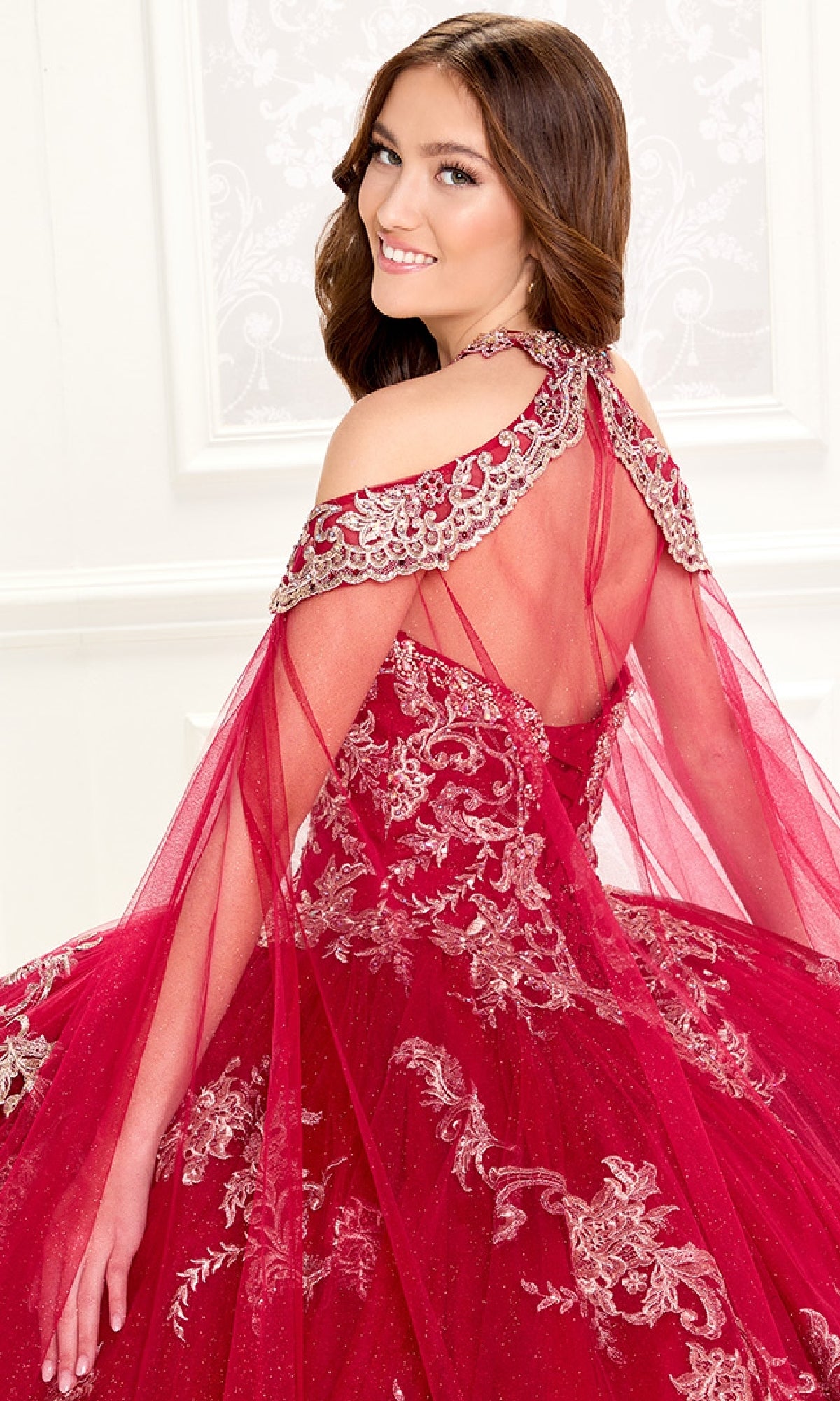 A young woman smiles as she poses with her back partly turned, wearing the Quinceanera Dress PR30086 by Princesa—a red, embroidered lace gown featuring sheer, beaded shoulder details.