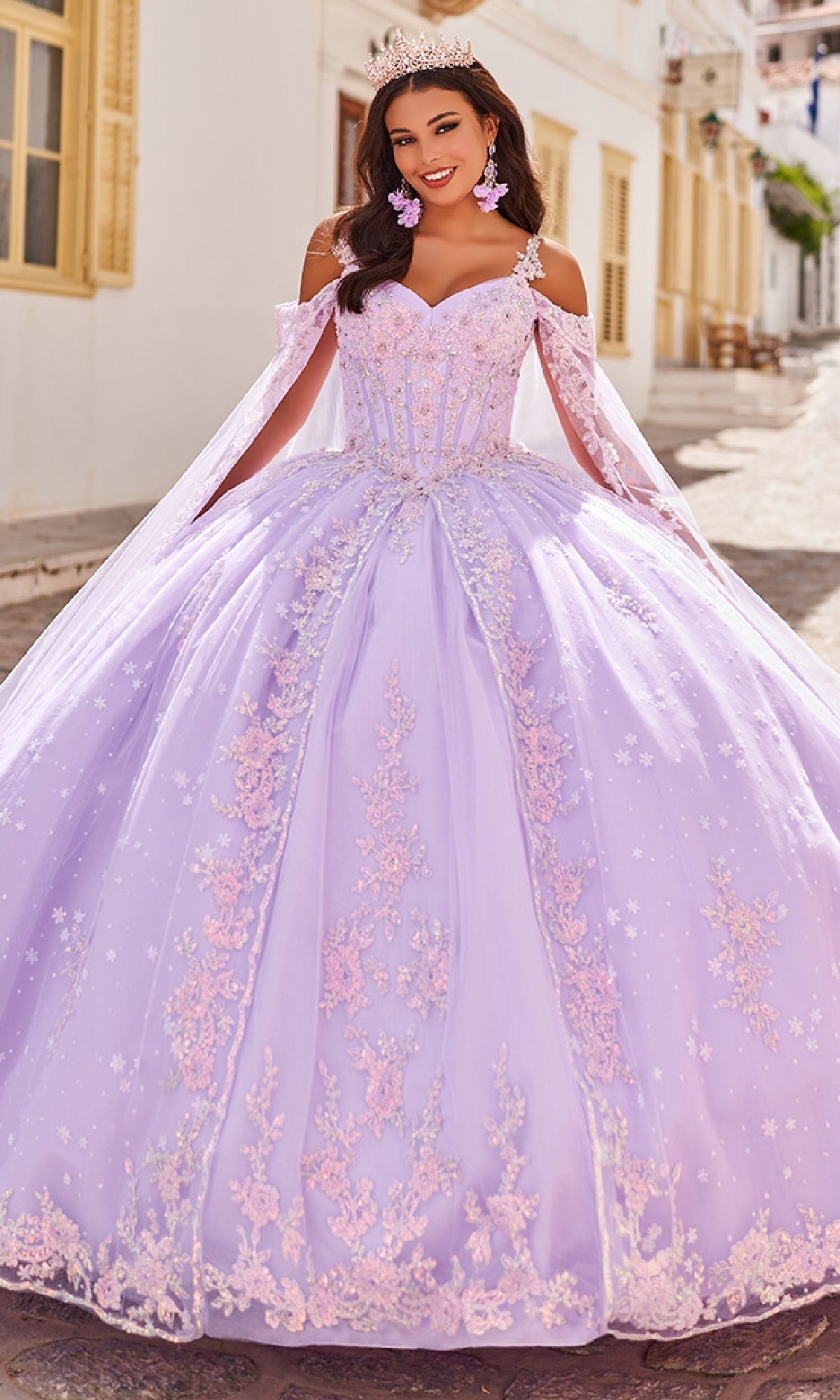 A woman stands outdoors wearing the Quinceanera Dress PR30087 By Princesa, featuring a lavender hue, corset bodice, pink embroidery, off-shoulder sleeves, a tiara, and pink floral earrings.