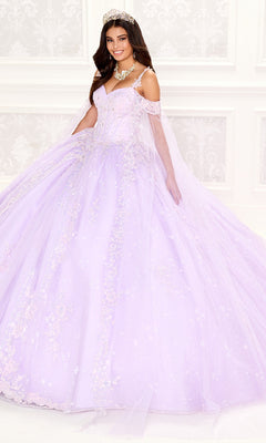 A woman wearing the Quinceanera Dress PR30087 By Princesa with floral details, a corset bodice, tiara, and jewelry smiles at the camera in front of a white ornate wall.