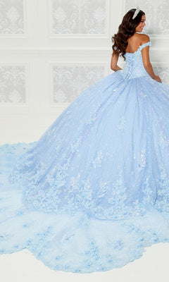 A woman wearing the Quinceanera Dress PR30111 By Princesa, an off-the-shoulder light blue gown with a cathedral train and lace embroidery, stands facing away in an elegant room with white patterned walls.