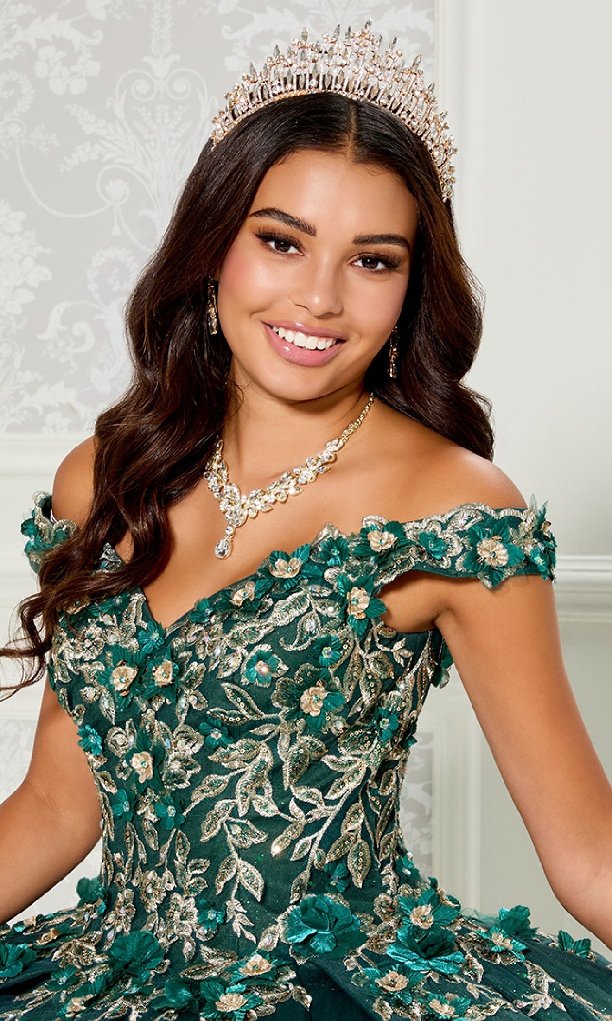 A young woman poses indoors in the Quinceanera Dress PR30114 By Princesa, a green and gold off-the-shoulder gown featuring 3D floral appliques, paired with a jeweled tiara and matching necklace against a light decorative background.