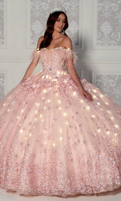 A woman wearing the Quinceanera Dress PR30115 By Princesa, featuring lights and floral accents, with a tiara, stands before an ornate wall. Her glowing pink off-the-shoulder gown highlights her elegant celebration.