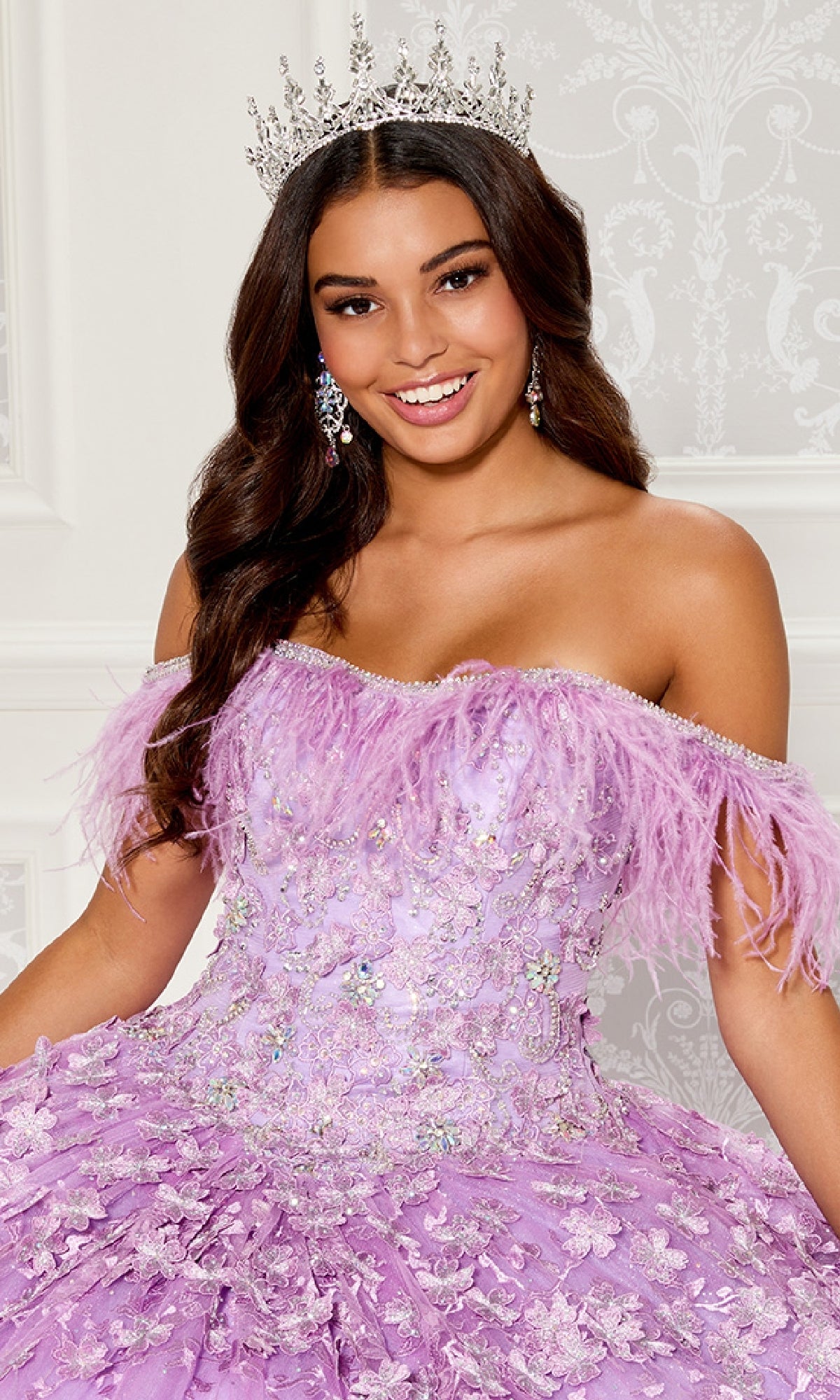 A woman smiles in a beautifully decorated room, wearing a silver crown and the Quinceanera Dress PR30115 By Princesa—an off-the-shoulder lavender gown with feather and sequin details.