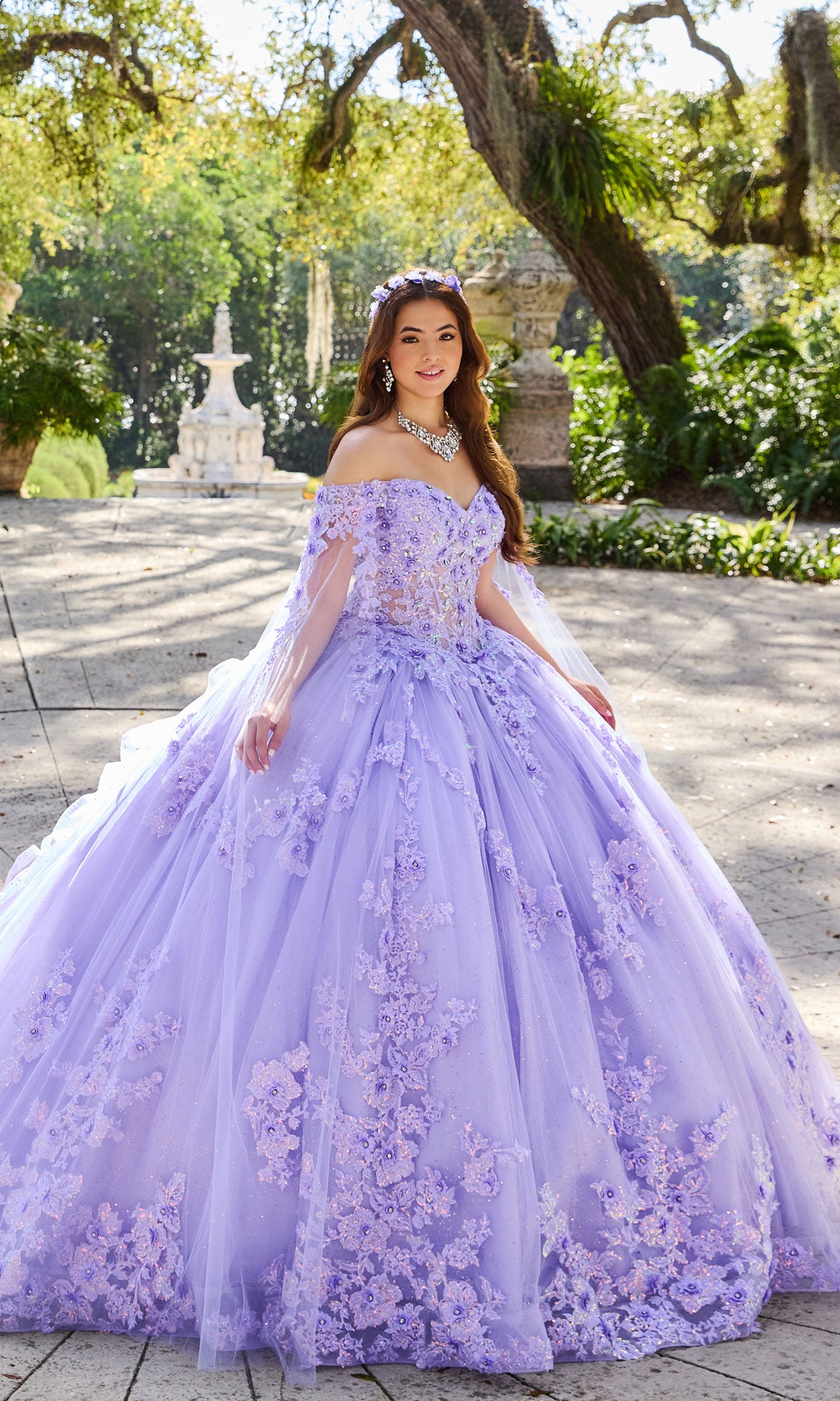 A woman stands outdoors in a garden wearing the Quinceanera Dress PR30116 By Princesa, featuring a lavender off-the-shoulder design with floral appliqués, a fitted corset bodice, and a sparkling necklace.