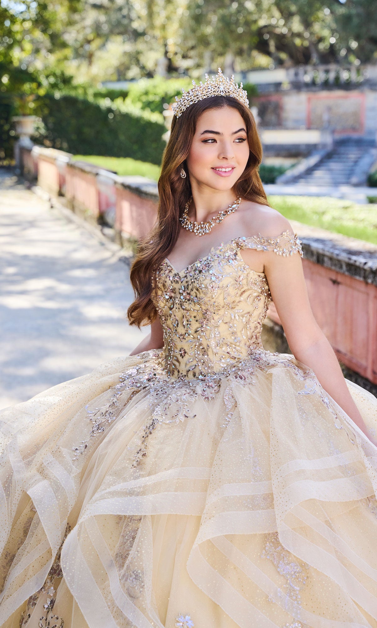 A young woman in the Quinceanera Dress PR30117 By Princesa, featuring a detachable cape and jeweled tiara, stands outdoors on a sunny day, smiling as she looks to the side.