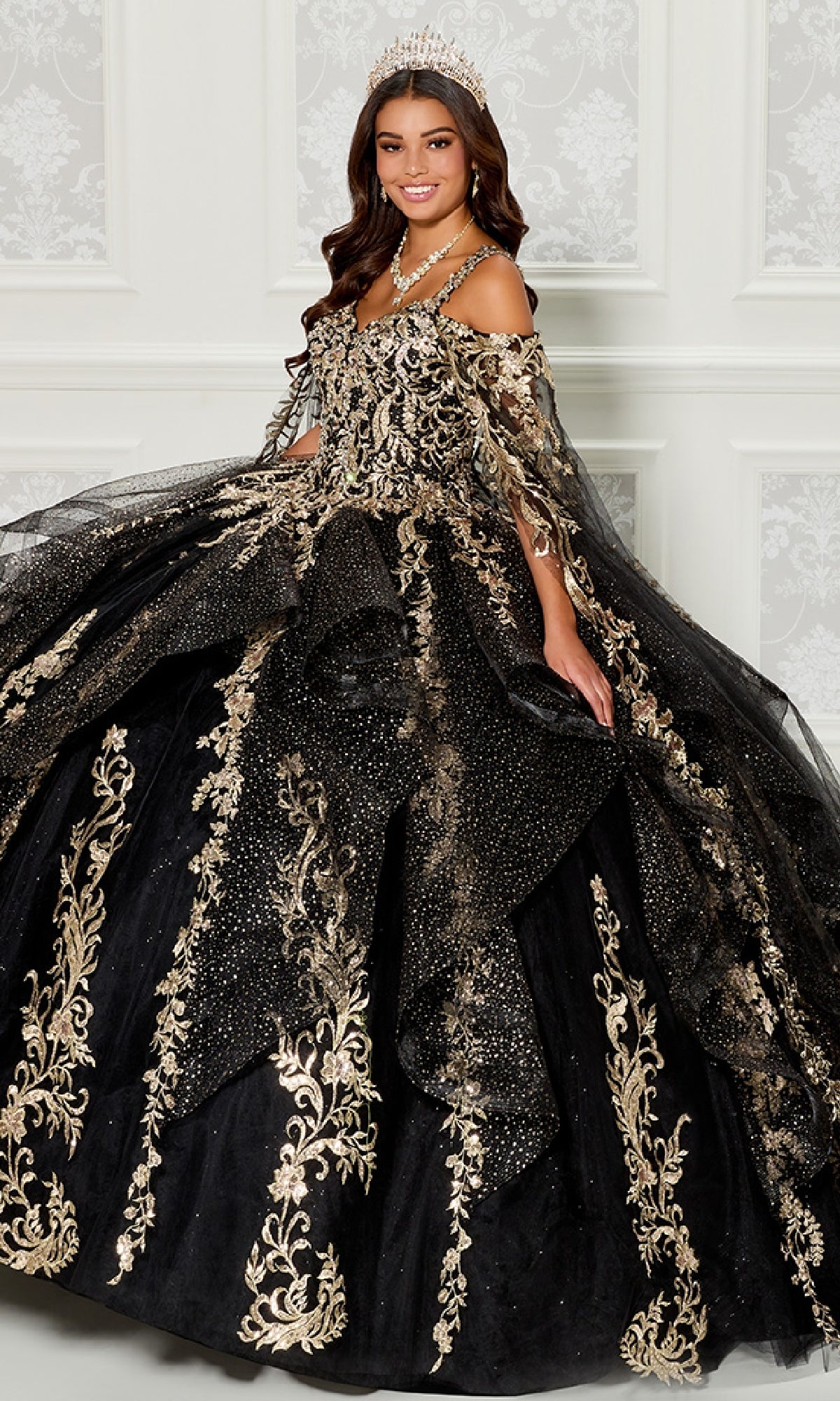 A woman wears the Quinceanera Dress PR30117 By Princesa—an elaborate black gown with gold embroidery, tiara, and a dramatic detachable cape—standing indoors against a white patterned wall for an elegant look.