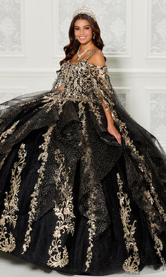 A woman wears the Quinceanera Dress PR30117 By Princesa—an elaborate black gown with gold embroidery, tiara, and a dramatic detachable cape—standing indoors against a white patterned wall for an elegant look.